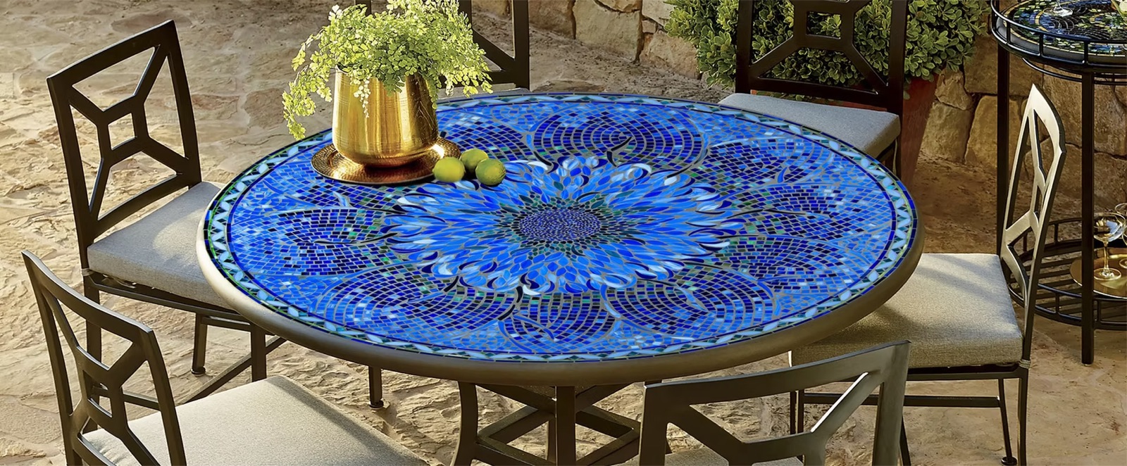 Round mosaic-top dining table in deep blues, set with iron chairs and a brass vase of greenery
