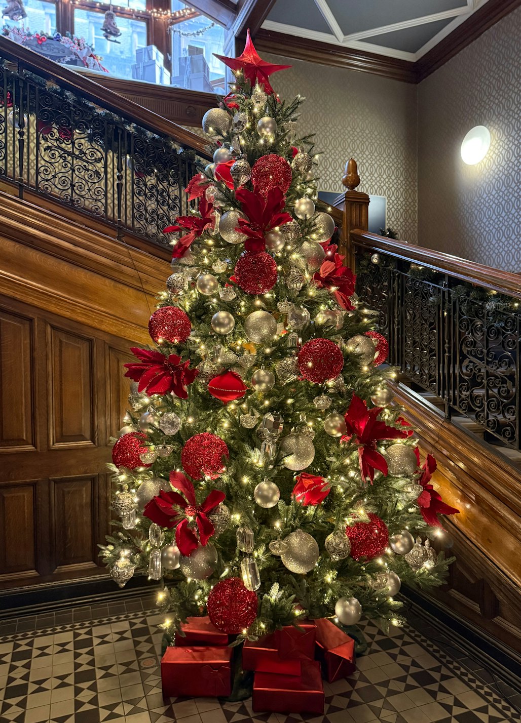 Christmas tree themed in red and silver, with big red bows and wrapped gifts, at the foot of a wooden staircase